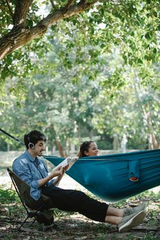 Side view of positive young multiracial couple spending time together in forest with book and headphones and resting in hammock