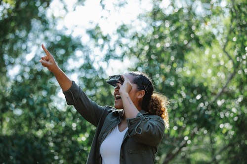 Happy young Hispanic female in casual wear looking at small binocular in green woods and pointing away