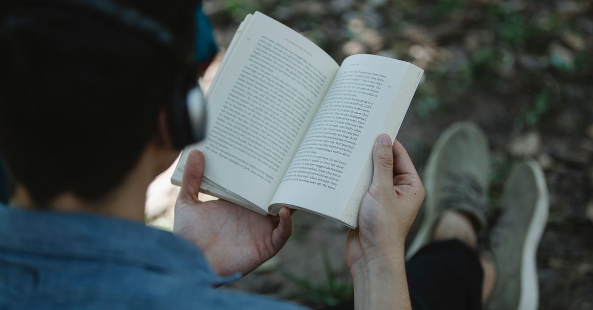 From above back view of crop unrecognizable male listening to music via headphones and reading book while sitting on cha