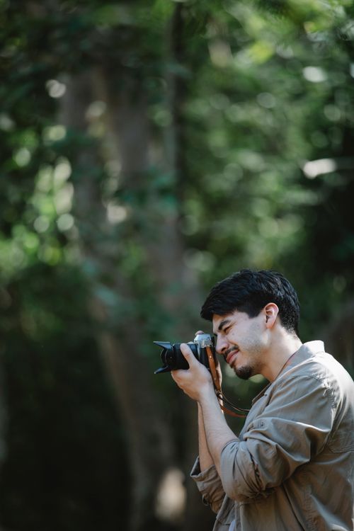 Free Side view of faceless concentrated man in casual outfit standing in green woods near trees while taking pictures on photo camera in sunny summer day Stock Photo