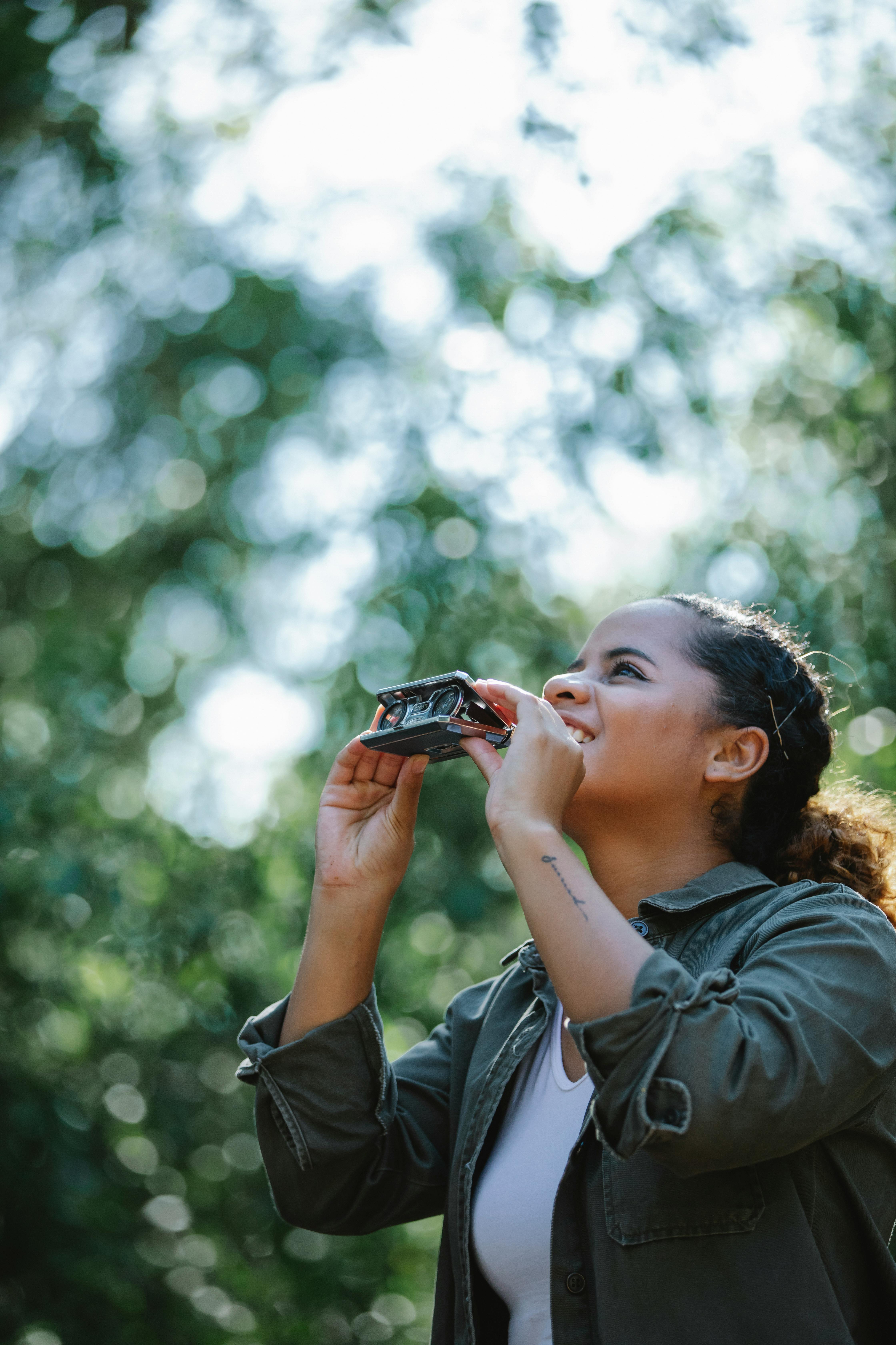 From below of young smiling Hispanic female in casual outfit with a small binocular in hands standing in forest and looking up