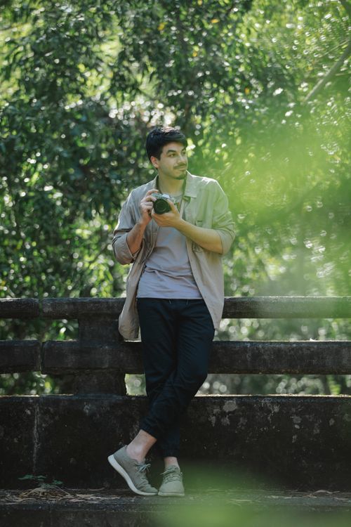 Free Full length young delighted male photographer wearing casual clothes standing near fence on footbridge with vintage photo camera and looking away Stock Photo