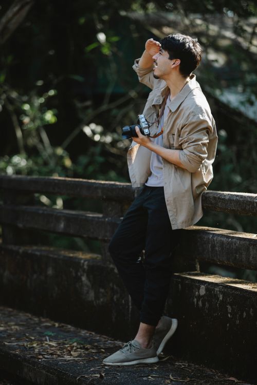 Free Full body young male photographer in casual wear standing near fence on wooden footbridge with retro photo camera and looking away Stock Photo
