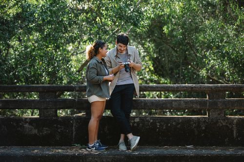Free Full body smiling diverse couple in casual wear with photo camera standing near fence on wooden footbridge in park Stock Photo