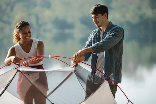 Young diverse traveling couple in casual outfits putting up tent near lake in summer day