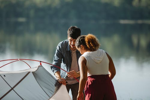 Focused young diverse couple of tourists putting up tent near pond in summer day