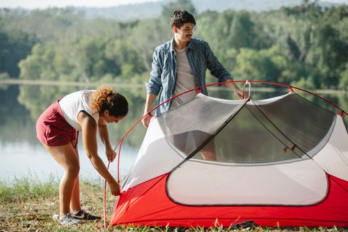 Free Full body young diverse couple of travelers putting up colorful tent on grassy river bank Stock Photo
