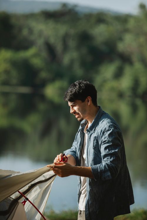 Free Side view of young bearded male traveler in casual wear putting up tent in campsite on river bank in summer day Stock Photo