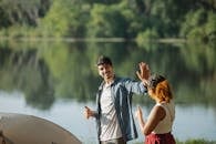 Happy young traveling couple giving each other high five while standing on river bank and putting up tent