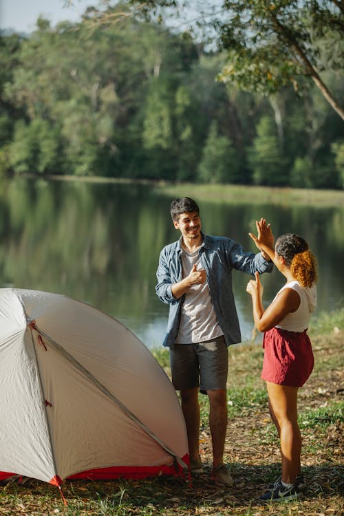 Content male tourist and anonymous ethnic female beloved giving high five with thumbs up while looking at each other on river coast