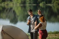 Glad male hiker with unrecognizable ethnic female beloved showing thumbs up and giving high five while looking at each other in campsite