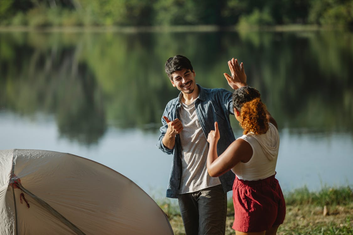 Free Glad male hiker with unrecognizable ethnic female beloved showing thumbs up and giving high five while looking at each other in campsite Stock Photo
