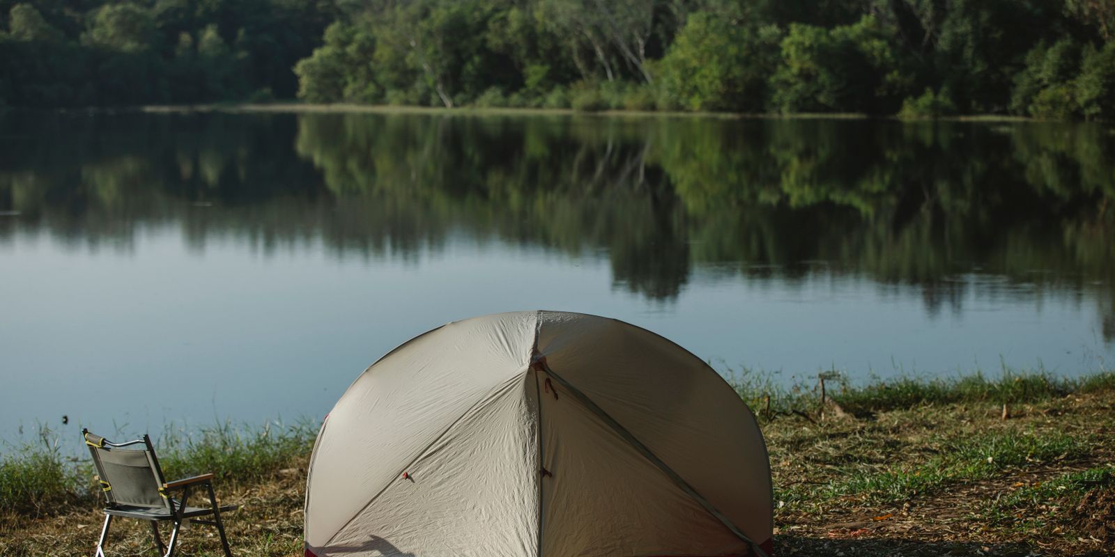 A tent and folding chair on a grassy shore beside still water