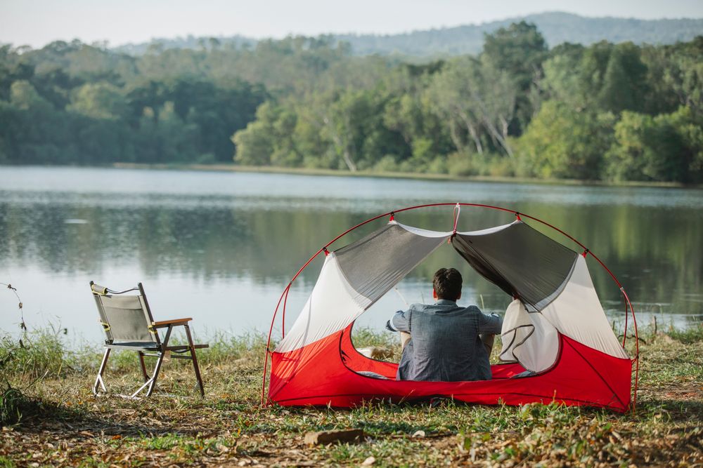 Camper sitting in a tent by a calm lake next to a camp chair