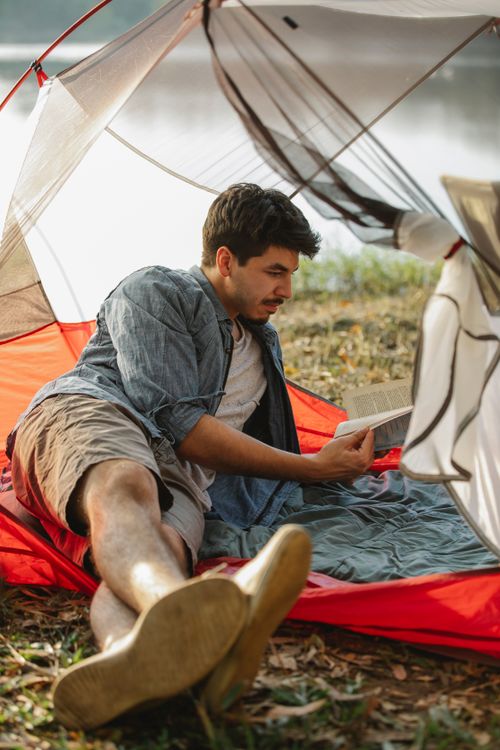 Free Adult male traveler with crossed legs reading textbook while lying in tent on lake coast in sunlight Stock Photo