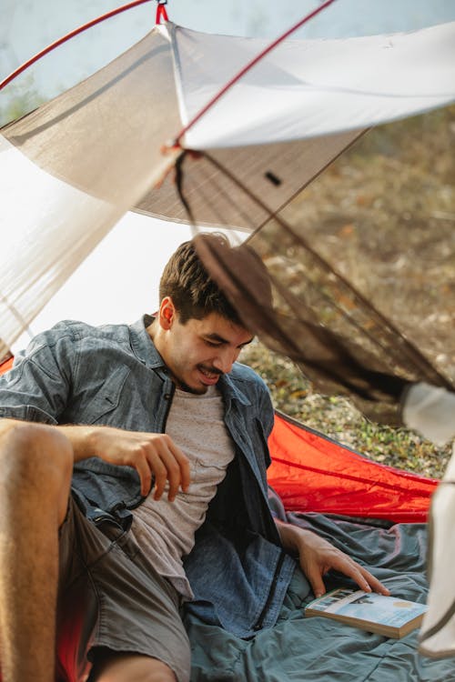 Adult unshaven male tourist with textbook resting in tent and looking down during summer trip on sunny day