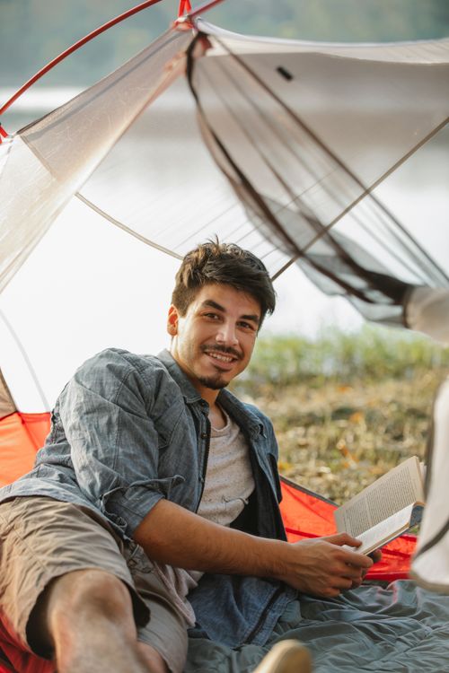 Free Adult content male hiker with textbook lying in tent against river and looking at camera Stock Photo
