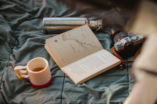 From above of open textbook between mug of hot drink and retro photo camera on crumpled fabric