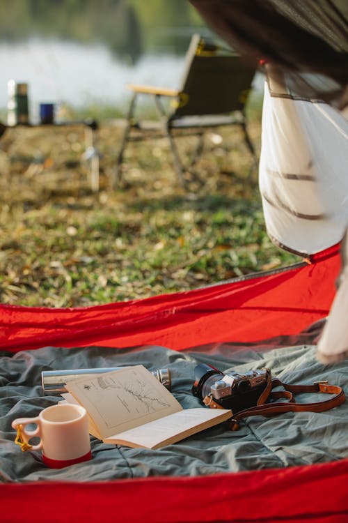 Hot drink in mug near open textbook on crumpled fabric in tent on lake shore