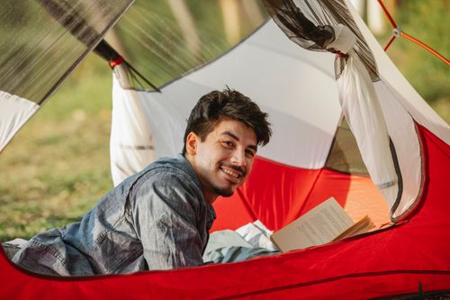 Free Side view of cheerful adult male hiker lying with textbook in tent and looking forward in sunlight Stock Photo