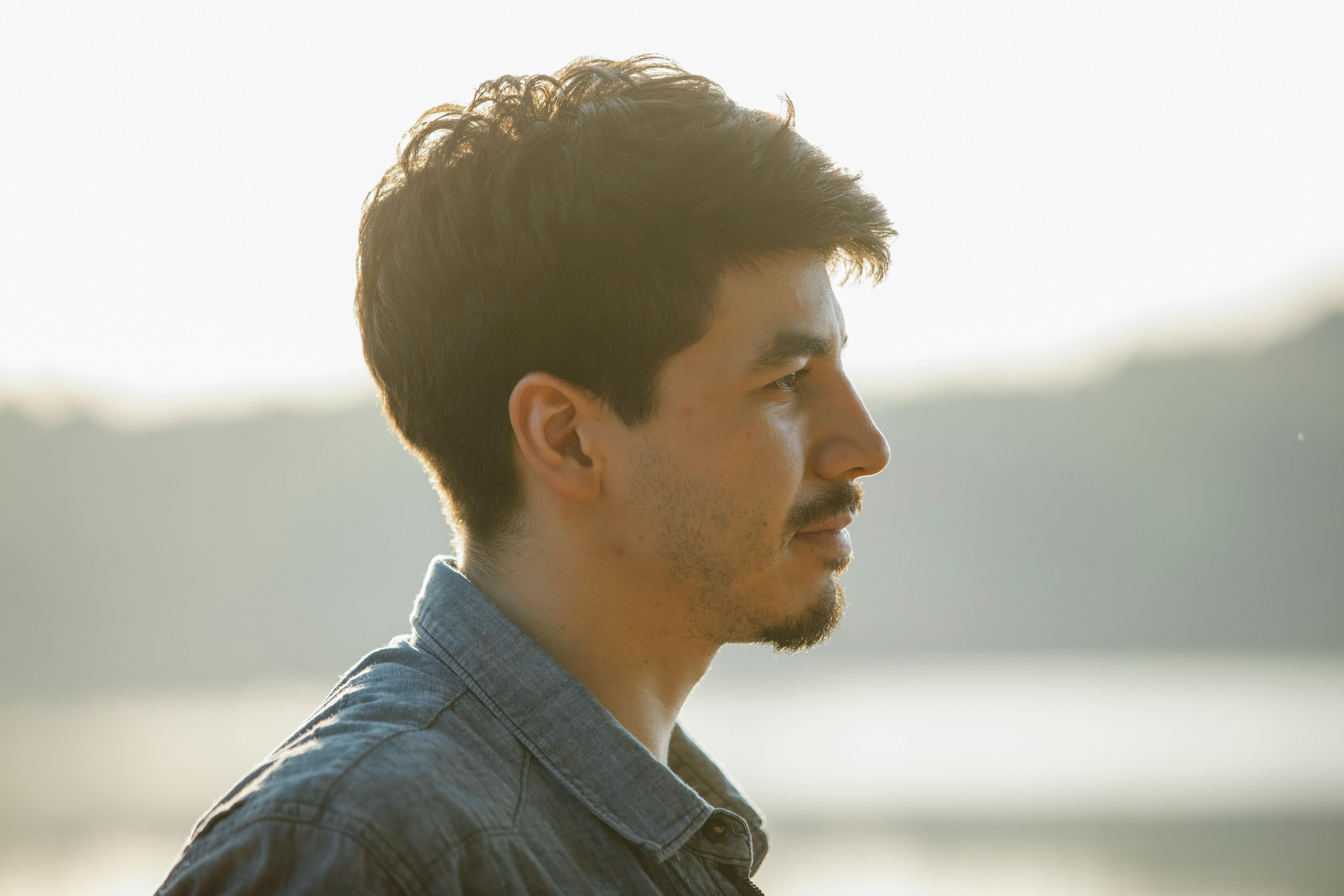 Profile of a young man looking pensive as he gazes over a serene lakeside setting outdoors.