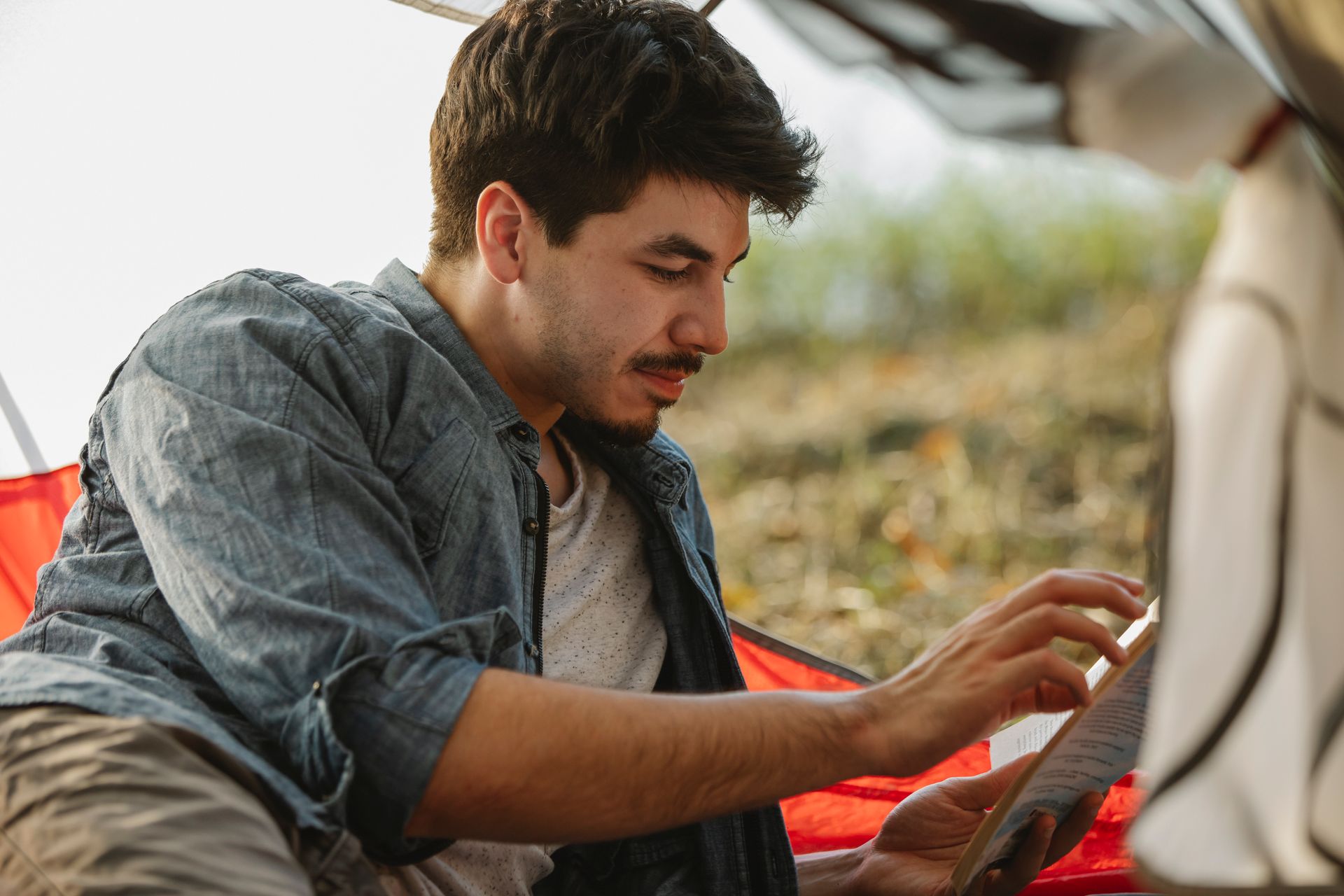 Crop focused male in casual wear reading book and lying in camp tent in peaceful nature