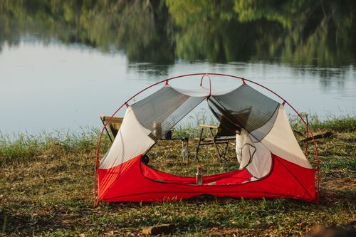 Free Camping tent and chairs placed on grassy shore of peaceful lake surrounded by lush green forest during romantic picnic on sunny day Stock Photo