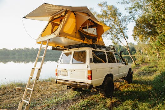 Off road car with camping tent on roof parked on grassy coast of picturesque lake surrounded by lush green trees against cloudless blue sky