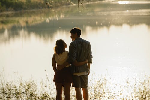 Free Back view of unrecognizable enamored young male and female travelers in casual clothes hugging and looking at each other while recreating on peaceful lake shore during romantic trip at sunset Stock Photo