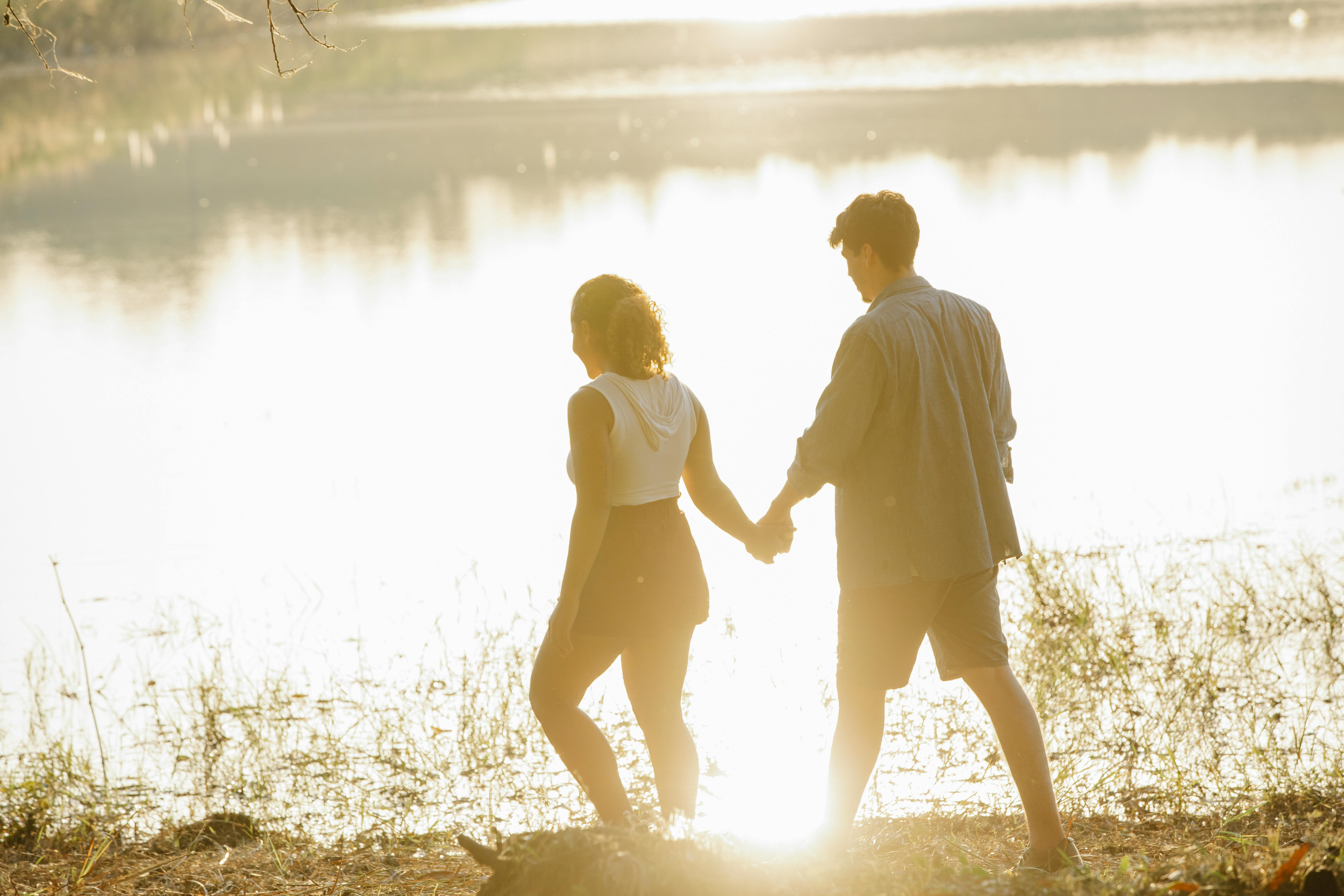 A couple holding hands and walking along a serene lakeside during sunset, capturing a romantic moment outdoors.