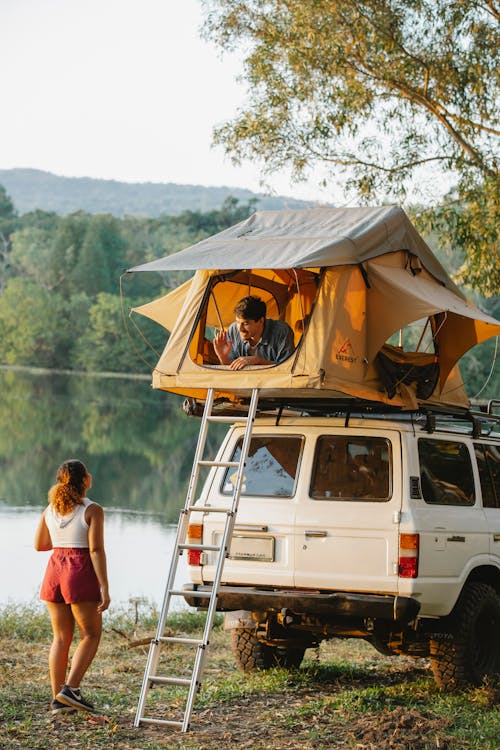 Free Back view of unrecognizable young female traveler standing on lake shore and talking to boyfriend resting in tent placed on SUV car roof Stock Photo