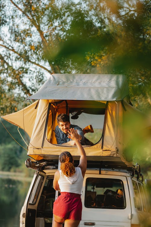 Back view of unrecognizable young female camper standing on ladder and giving high five to smiling boyfriend recreating in tent placed on SUV car roof at lakeside