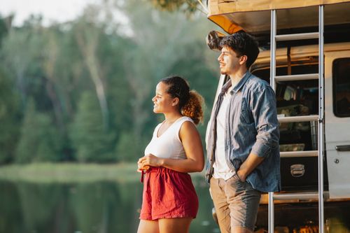 Free Side view of content young multiracial couple in casual outfits standing near SUV car on lake shore and admiring nature during road trip in nature at sunset Stock Photo