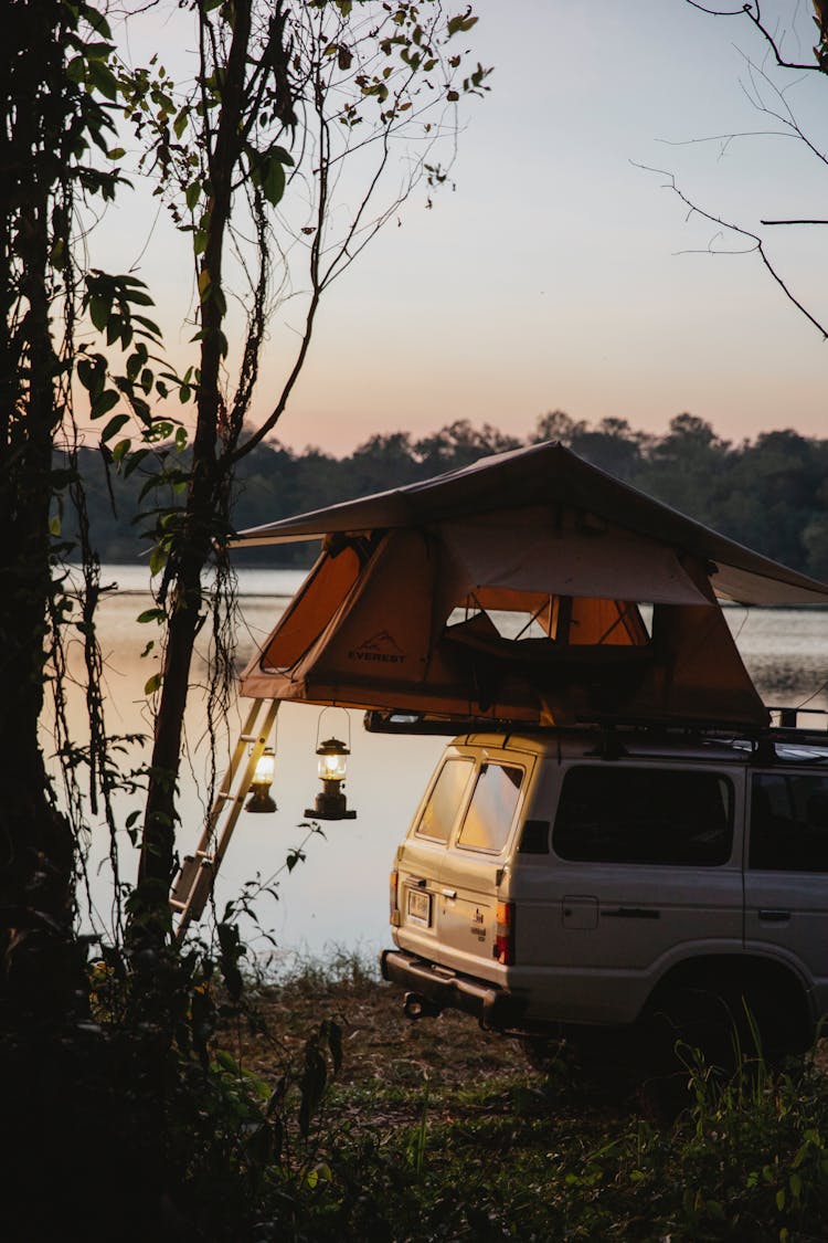 SUV Car With Camping Tent On Roof Parked At Lakeside Against Sundown Sky