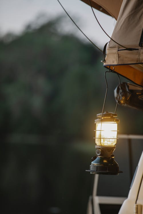 Glowing vintage lamp hanging from camping tent placed on roof of car parked on lake shore reflecting lush green forest at sundown