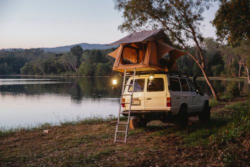 Free Camping tent with glowing retro lamps placed on top of white off road vehicle parked on grassy shore on peaceful lake surrounded by lush green tress against cloudless evening sky Stock Photo