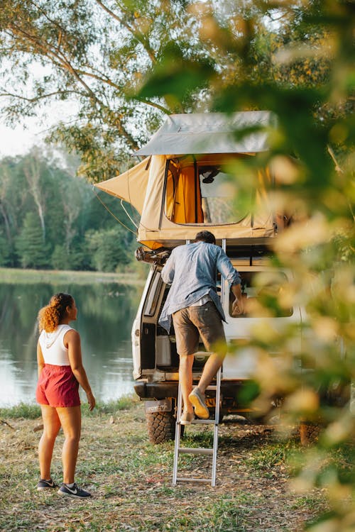 Back view of unrecognizable young male camper going downstairs from tent placed on car roof parked near lake during romantic trip with girlfriend