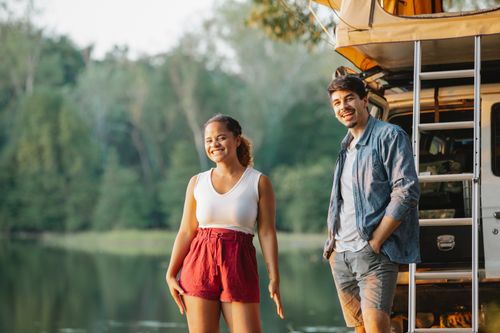 Free Excited young diverse couple in casual clothes smiling and looking at camera while recreating on lake shore standing near car during camping in forest on sunny day Stock Photo