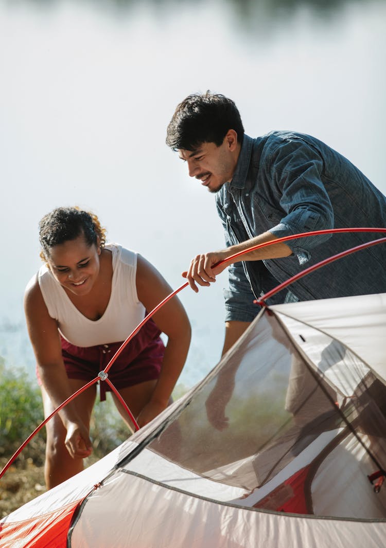 Happy Young Multiracial Couple Setting Up Tent On Lake Shore After Trekking