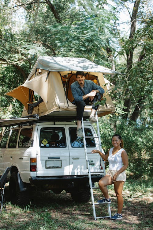 Full body of cheerful young diverse couple in casual clothes smiling while chilling together near SUV car with tent on top during camping in forest on sunny day