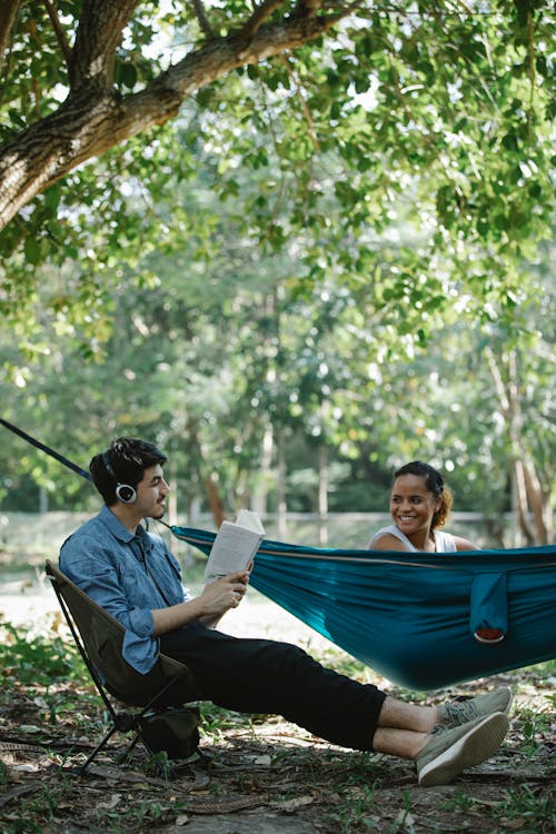 Positive male in headphones sitting with book on chair and looking on happy Hispanic girlfriend sitting on ground among green trees in park in sunny day