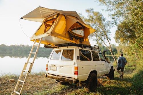 Back view of full body male traveler near yellow camping tent placed on roof of white car parked on coast of pond at daytime