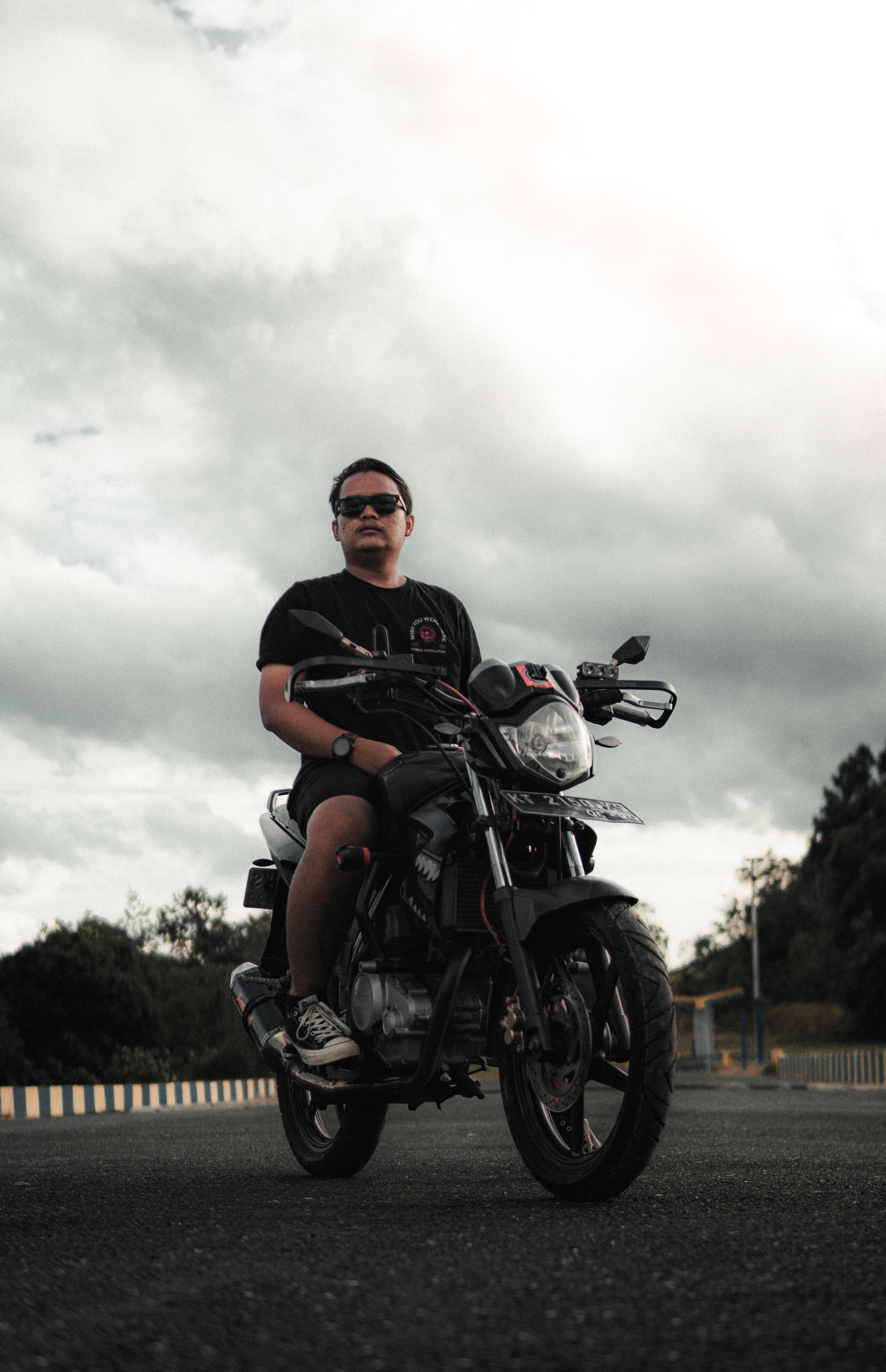 A man wearing sunglasses rides a motorcycle on an empty road beneath a cloudy sky.