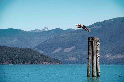 Free An adventurous man diving from a tall wooden post into a tranquil lake surrounded by mountains. Stock Photo
