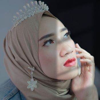 Charming Islamic female with stylish makeup wearing traditional hijab decorated with diadem and brooch touching face tenderly and looking away dreamily