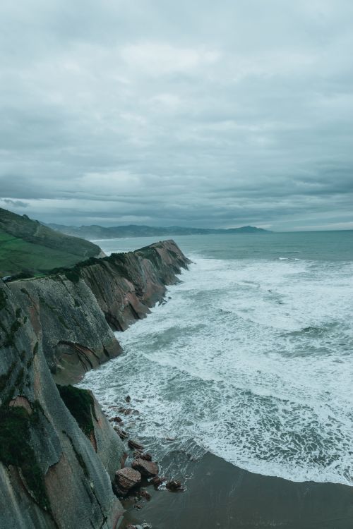 Free From above of powerful ocean with big foamy waves washing wet sandy beach surrounded by rocky cliffs against overcast sky in Gaztelugatxe Stock Photo