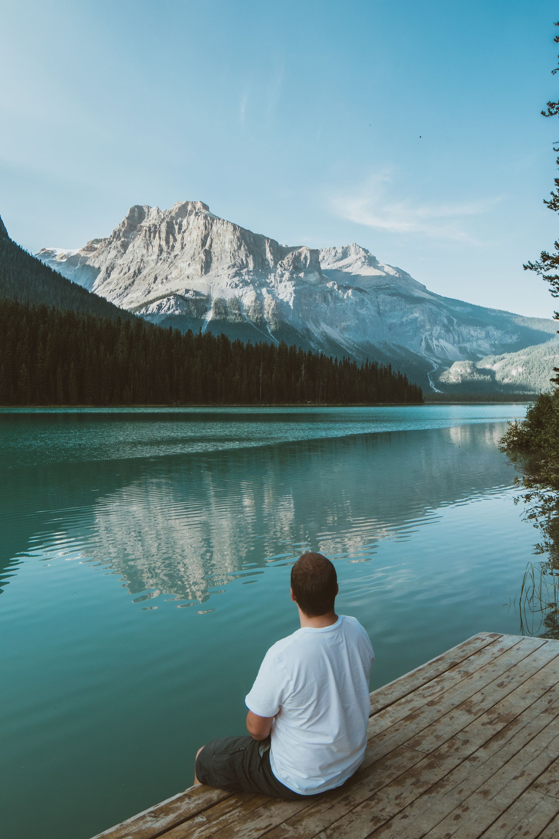 A man in a white T-shirt sitting on a wooden dock, looking across a turquoise mountain lake toward a snow-capped peak on a clear day