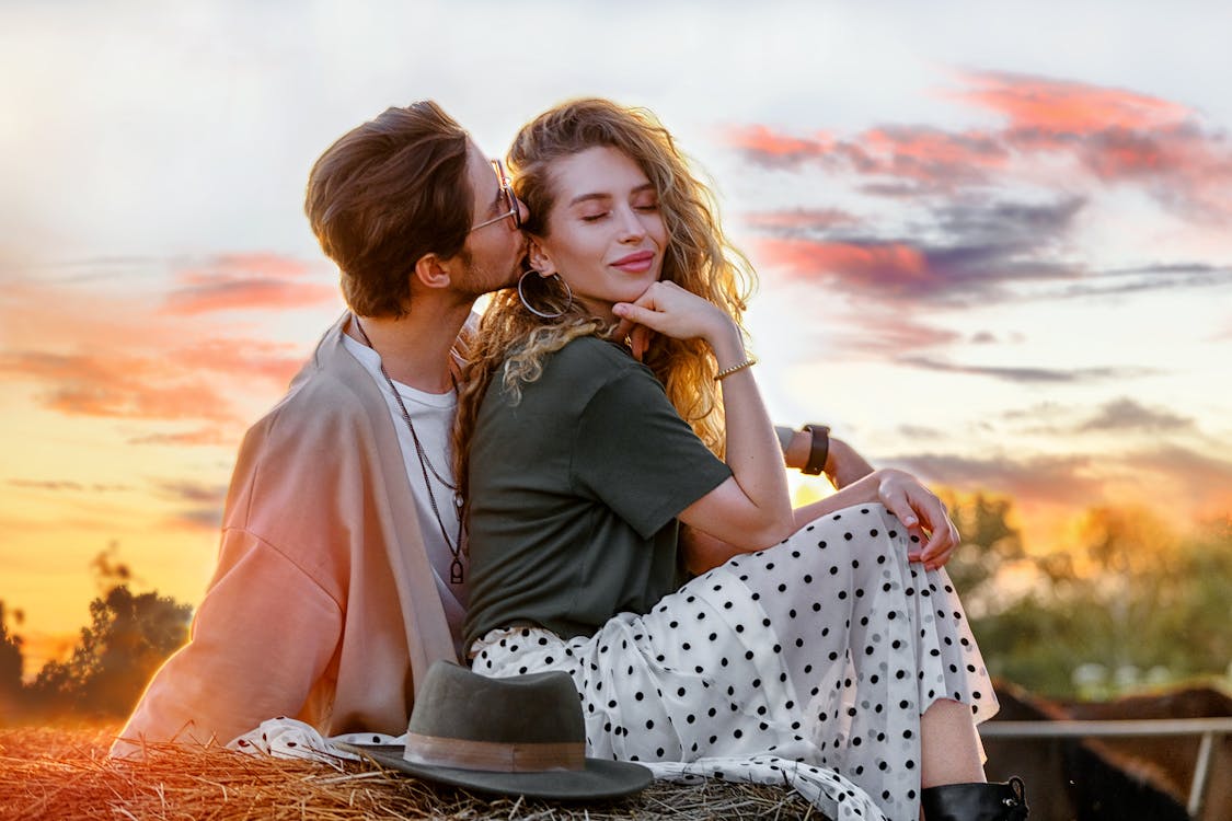 Free Side view serene young couple in stylish wear kissing and bonding while sitting on haymow against picturesque sunset sky Stock Photo