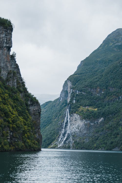 Magnificent landscape of wide river with rippling water surface flowing near massive rocky formations covered with green plants under overcast sky