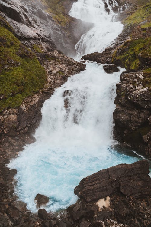 Foamy waterfall falling down rocks and stones covered with green moss in mountainous terrain